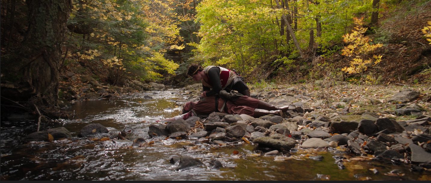 Edmund kneels over a fallen body in a forest stream